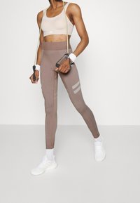 Jasnobrązowe dopasowane legginsy z bocznymi paseczkami, zestawione z beżowym stanikem sportowym. Dodatki to gumy oporowe i białe sneakersy.