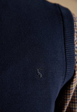 Dunkelblaue Strickware mit einem kleinen gestickten Hasenlogo, geschichtet über einem karierten Hemd, das am Rand sichtbar ist.