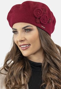 Berretto di lana rosso con decorazioni floreali, featuring tre rose di stoffa e dettagli a spirale. Texture liscia, forma rotonda e vestibilità comoda.