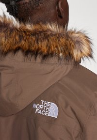 Brun hettejakke med faux fur-kant og brodert hvitt logo. Tekstuert stoff med synlige sømmer og et rent design.