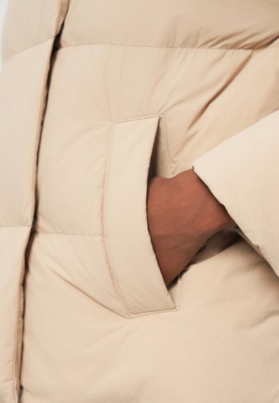 Beige puffer jacket fabric with a smooth texture. Features a side pocket with a hand tucked in and a partially visible sleeve edge.