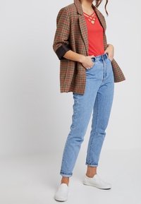 Geruite blazer in bruin en rode tinten, rode top met kruislings decolleté, hoog getailleerde blauwe jeans en witte instapschoenen.