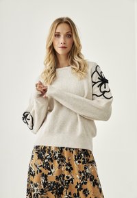 Suéter de punto crema con bordado floral negro en las mangas, combinado con una falda de estampado floral en tonos de negro y mostaza.