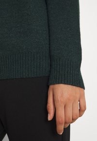 Pull vert foncé avec un détail de poignet côtelé, fabriqué dans une texture douce. Le poignet dépasse légèrement le poignet. Tissu noir visible en dessous.