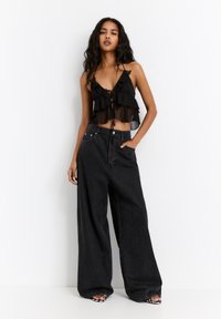 Black sheer crop top with layered ruffles and tie front, paired with high-waisted wide-leg black jeans. Model stands with one hand on hip.