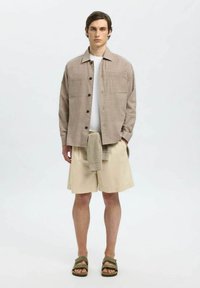 Stribet skjorte i beige nuancer, beige shorts og olivengrønne sandaler; har knaplukning, to brystlommer og en foldet sweater om livet.