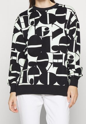 Sudadera en blanco y negro con patrón abstracto de letras grandes en bloque, usada con pantalones blancos, mostrada desde el cuello hasta las caderas.