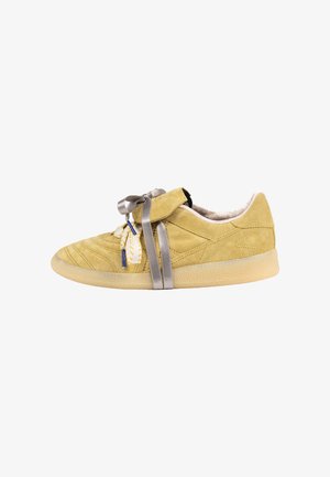 Suede low-top sneaker in mustard yellow, featuring a beige rubber sole, decorative tie in gray satin, and textured stitching detail on the upper.