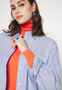 Camisa azul clara com riscas, sobre uma blusa de gola alta laranja brilhante, com anéis e brincos coloridos. Close-up das mãos e do rosto.