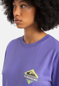 Paarse katoenen t-shirt met een ronde halslijn. Heeft een kleurrijk logo met geometrische vormen in het geel, groen en blauw op de borst.