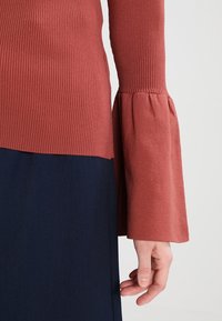 Gestreiftes, rostfarbenes Langarmoberteil mit schwingenden Ärmelabschlüssen, kombiniert mit einem dunkelblauen Unterteil. Verfügt über einen lockeren Schnitt und eine weiche Textur.