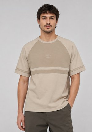 Uomo con capelli scuri che indossa una camicia beige e marrone chiaro a maniche corte con logo Umbro, in piedi davanti a uno sfondo chiaro e uniforme.