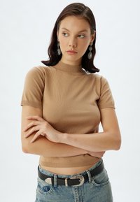 Lysebrune ribbede kortærmede top med høj krave, parret med højtaljede blå denim jeans og et sort bælte med en sølvspænde.
