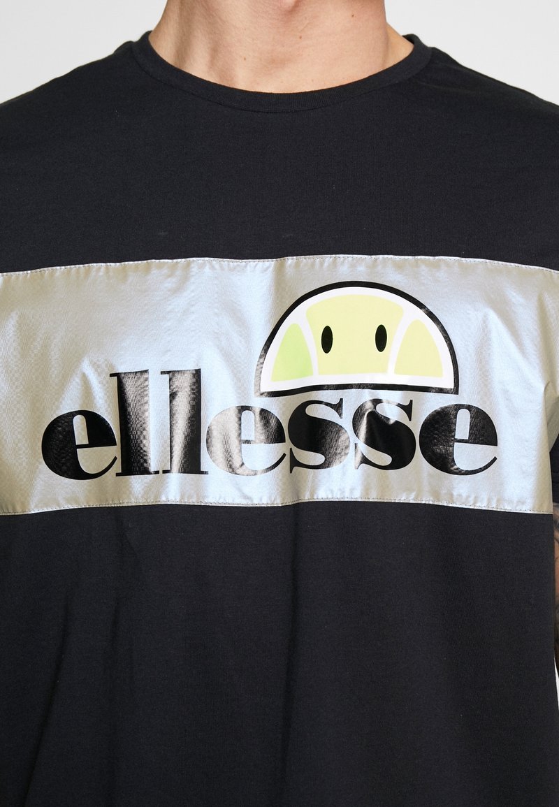 Chemise noire avec une large bande horizontale argentée arborant le logo "ellesse" en noir, surmonté d'un design semi-circulaire de balle de tennis au-dessus du texte.