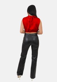 Donna con lunghi capelli scuri che indossa un top rosso senza maniche annodato al collo, pantaloni in pelle nera e scarpe con tacco alto bianche, di spalle.