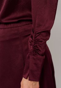 Bordeaux blouse van textuurstof, met geraffineerde mouwen en een manchetontwerp. Gladde oppervlakte met een subtiel bladdessin.