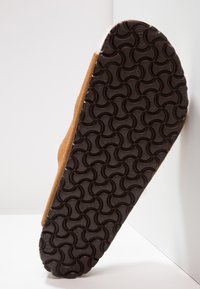 Bruna mockaskor som visar en texturerad brun gummisula med ett vågigt mönster, sett från botten i en vinkel mot en ljus bakgrund.