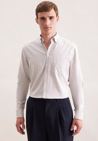 Camicia bianca con un piccolo motivo a quadretti blu e marrone, abbinata a pantaloni blu scuro. La camicia ha maniche lunghe e colletto.