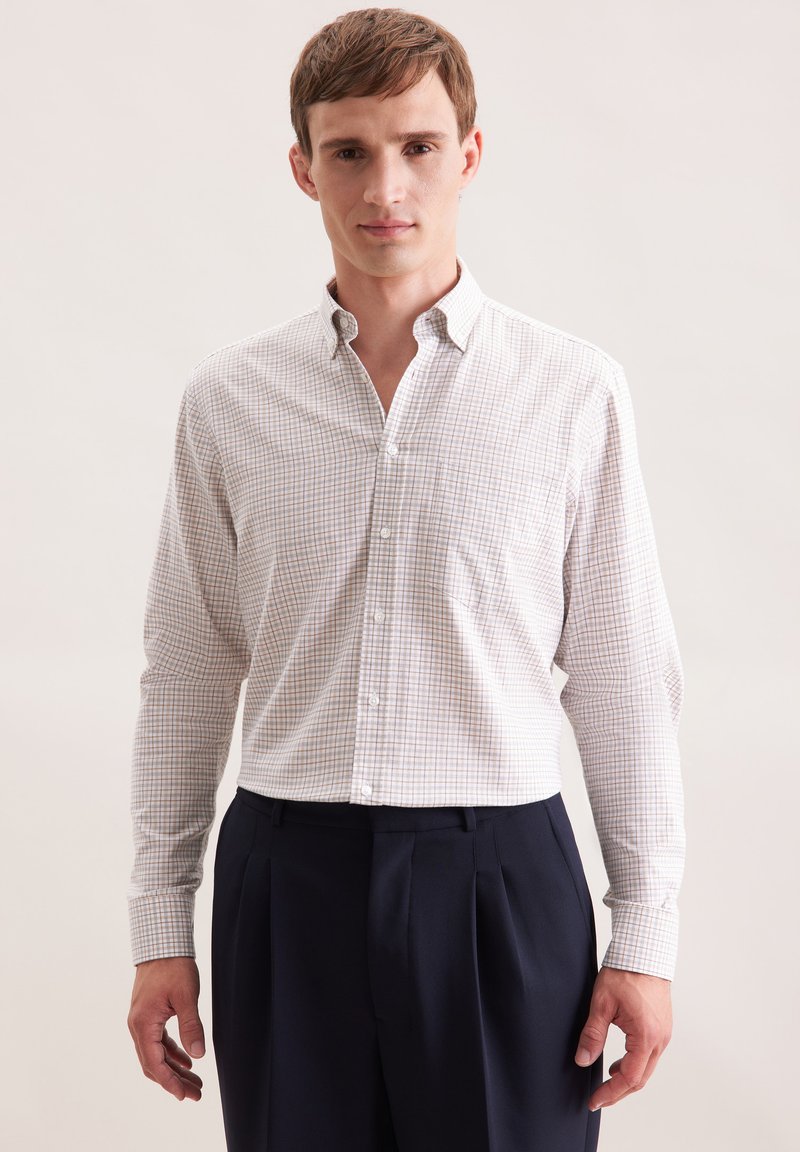 Camicia bianca con un piccolo motivo a quadretti blu e marrone, abbinata a pantaloni blu scuro. La camicia ha maniche lunghe e colletto.