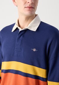 Marineblå bomull rugby-shirt med en kremfarget krage, med horisontale striper i gul og oransje, og en GANT-logo på brystet.