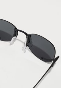 Ochelari de soare negri cu lentile ovale, sticlă întunecată, ramă subțire din metal negru și suporturi pentru nas transparente. Design elegant cu un hardware minimalist.