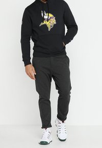 Felpa nera con il logo dei Vikings sul petto, abbinata a pantaloni felpati grigio scuro a gamba affusolata e sneakers bianche con dettagli verdi.