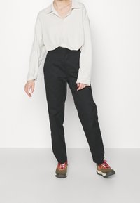Chemise boutonnée crème ample, pantalon noir taille haute et baskets brunes avec des lacets rouges. Design simple et couleurs neutres.