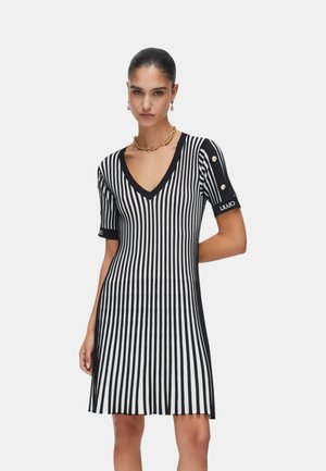 Donna che indossa un vestito a righe verticali bianche e nere con scollo a V, maniche corte, bottoni dorati sulle maniche e una collana a catena dorata.