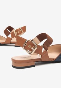 Sandalias planas marrones con una correa ancha, hebilla dorada y acentos elásticos. El tacón tiene un diseño de bloque bajo y un acabado texturizado.
