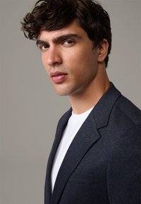 Granatowa marynarka o fakturowanej tkaninie, z wyraźnymi klapami i małym emblematem na lewym kieszonkowym piersiowym, noszona na białej koszuli.