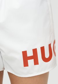 Λευκό σορτς με μεγάλους κόκκινους γράμματα «HUG» τυπωμένα κοντά στο τελείωμα, φορεμένα στο πάνω μέρος του μηρού ενός ατόμου.