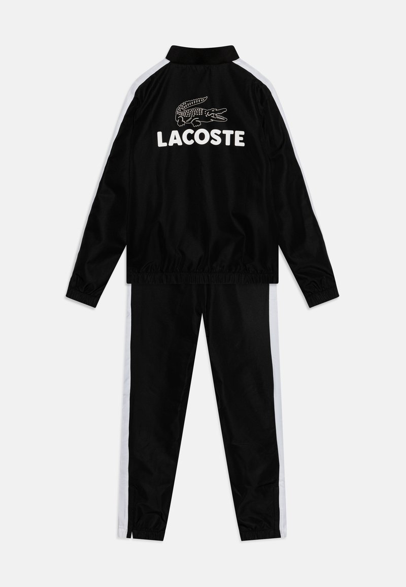 Survêtement de sport noir en tissu lisse, avec une bande blanche sur les côtés et un logo Lacoste bien en vue avec un graphique de crocodile.