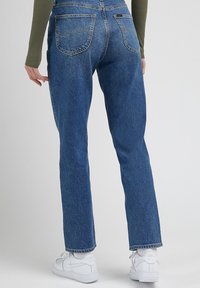 Modré džínové lacláče se spadlým střihem, vybavené zadními kapsami a opotřebovanou texturou, kombinované s bílými teniskami a zeleným tričkem s vroubkováním.