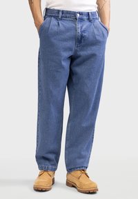 Pantalon en denim bleu avec une coupe décontractée, présentant des plis, des poches latérales et une fermeture à bouton. Assorti à des bottes en cuir tan à lacets.