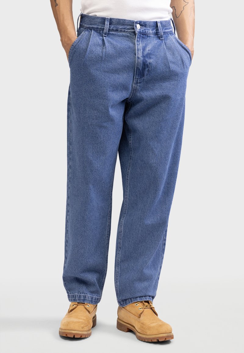 Pantalon en denim bleu avec une coupe décontractée, présentant des plis, des poches latérales et une fermeture à bouton. Assorti à des bottes en cuir tan à lacets.