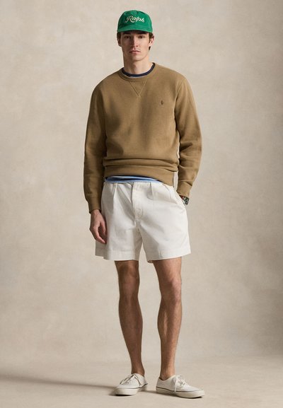 Νέος άνδρας που φοράει πράσινο καπέλο, καφέ φούτερ, λευκό κοντό φούστα με πιέτες και λευκά αθλητικά παπούτσια, στέκεται μπροστά σε ουδέτερο φόντο.