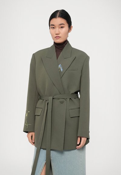 Blazer avvolgente verde oliva in tessuto strutturato, con due tasche frontali e ricami floreali, abbinato a un dolcevita marrone aderente e a una gonna in denim.