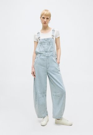 Ženska s kratkimi blond lasmi, oblečena v belo majico in široke svetlo modre jeans hlačne naramnice z rokami v žepih, stoji na belem ozadju.