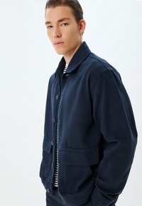Giacca blu navy in cotone con vestibilità rilassata, chiusura a bottoni, due tasche frontali e colletto semplice. Camicia a righe sotto.