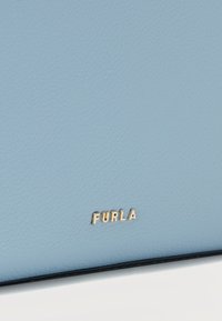 Επιφάνεια από δέρμα σε ανοιχτό μπλε με υφή, με μικρά χρυσαφένια μεταλλικά γράμματα που σχηματίζουν τη λέξη "FURLA" κοντά στο κάτω άκρο.