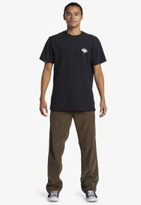 Black cotton t-shirt with a small white graphic on the chest, paired with green corduroy pants and black sneakers.