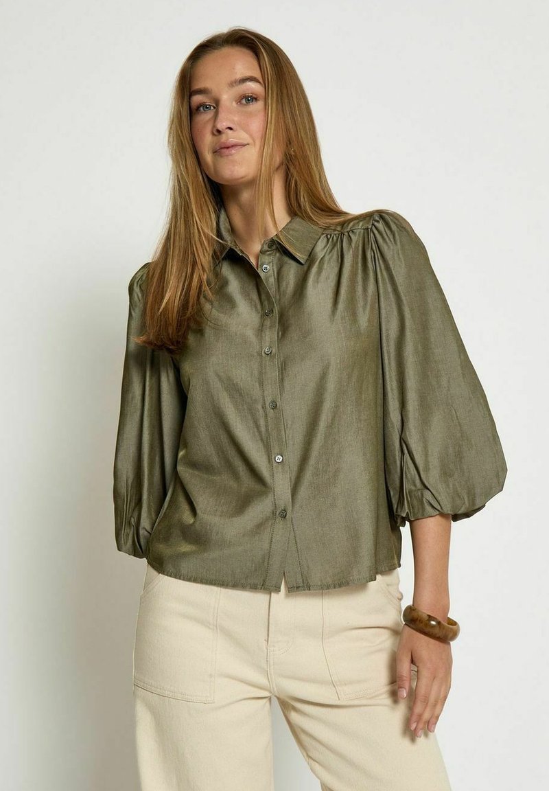 Olive green button-up blouse with puff sleeves, soft texture, and a straight fit. Paired with cream-colored pants, featuring a subtle sheen.