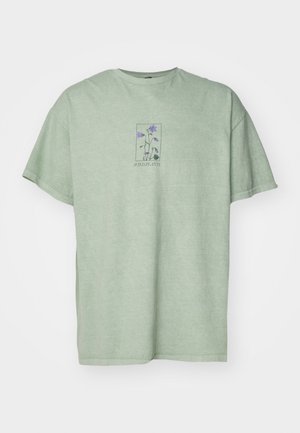 Lys grøn bomulds t-shirt med en løs pasform, der har et grafisk motiv af vilde planter i lilla nuancer inden for en rektangulær ramme.