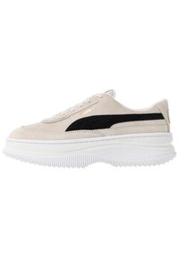 Beige mokkasneakers med en sort mokkastripe, hvit gummiplattformsåle og gullbranding. Har snøring og rund tå.