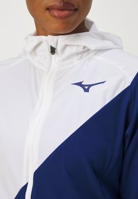 Balta un tumši zila jaka ar kapuci, rāvējslēdzēja aizdari un logo uz krūtīm. Materiāls izskatās gluds un ar vieglu tekstūru.