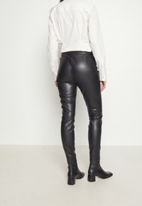 Personne portant un pantalon en cuir noir avec des talons et une chemise blanche à manches longues, debout contre un fond clair uni, vue de dos.