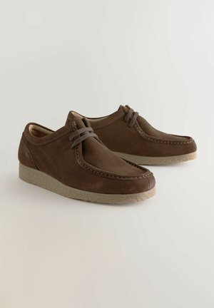 Zapatos de ante marrón con punta redondeada, diseño con cordones y suela de goma texturizada. Presentan detalles de costura a lo largo de la parte superior.