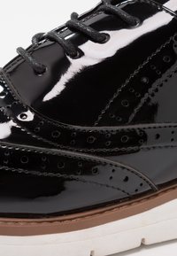 Chaussure brogue en cuir noir brillant avec détails perforés, lacets noirs et semelle épaisse beige et blanche.