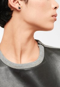 Boucles d'oreilles en triangle noir portées avec un t-shirt gris à col rond. Les boucles ont une finition mate et un design géométrique minimaliste.
