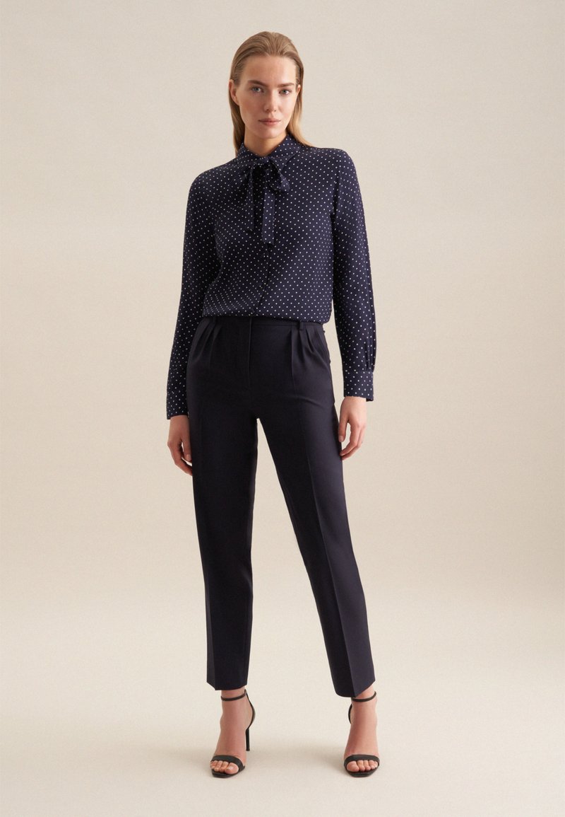 Blusa a pois blu navy con colletto annodato, abbinata a pantaloni neri su misura e sandali neri con cinturino, su uno sfondo neutro.